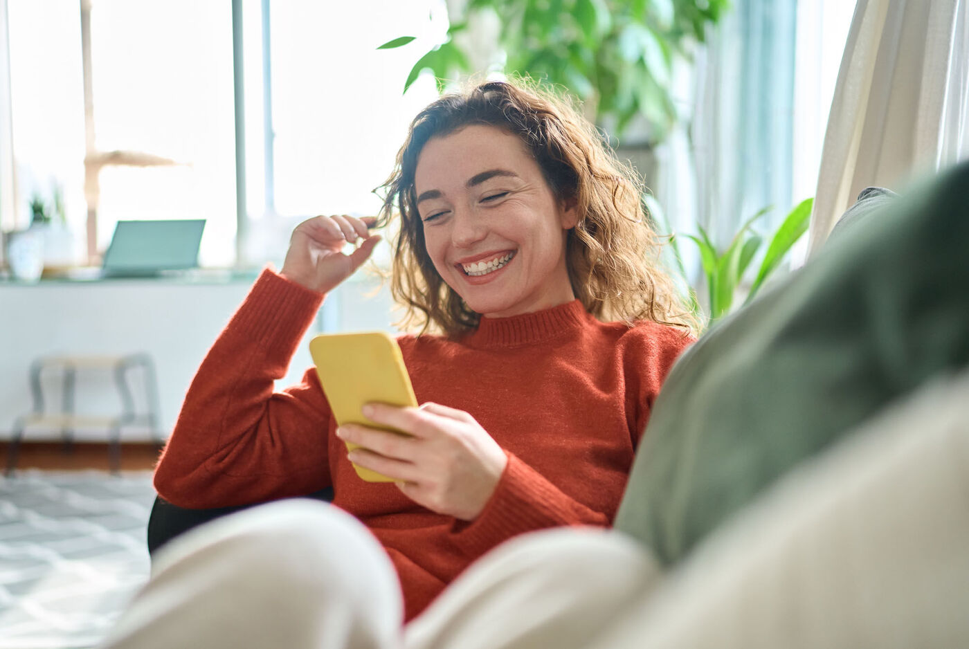 Una persona si rilassa su un divano guardando il suo smartphone in una stanza accogliente. Piante e un laptop sono sullo sfondo.